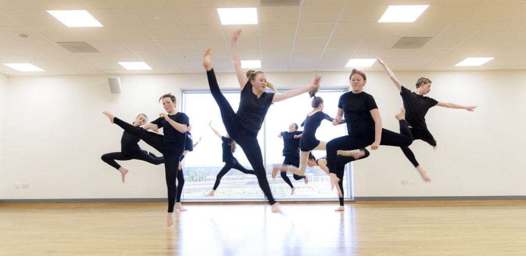 Active Schools Dance Classes in Elgin / Fochabers / Forres.10 young dancers leap in the air in a dance studio with a pale wood floor. There is a white wall behind them with a large window in the centre. All dancers are wearing black.