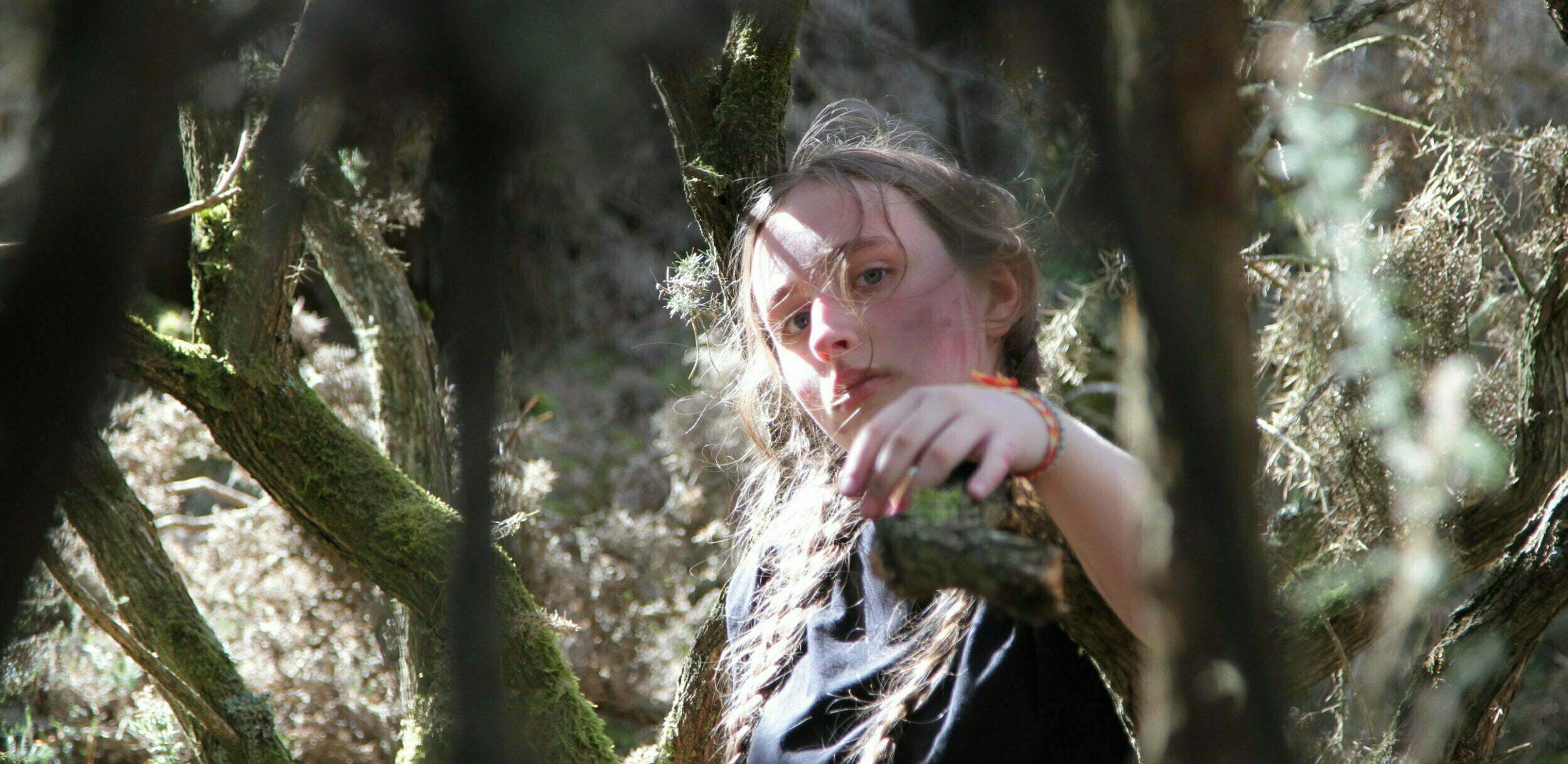 A young dancer in a black t-shirt stands in the woods amid gnarled trees and reaches toward the camera