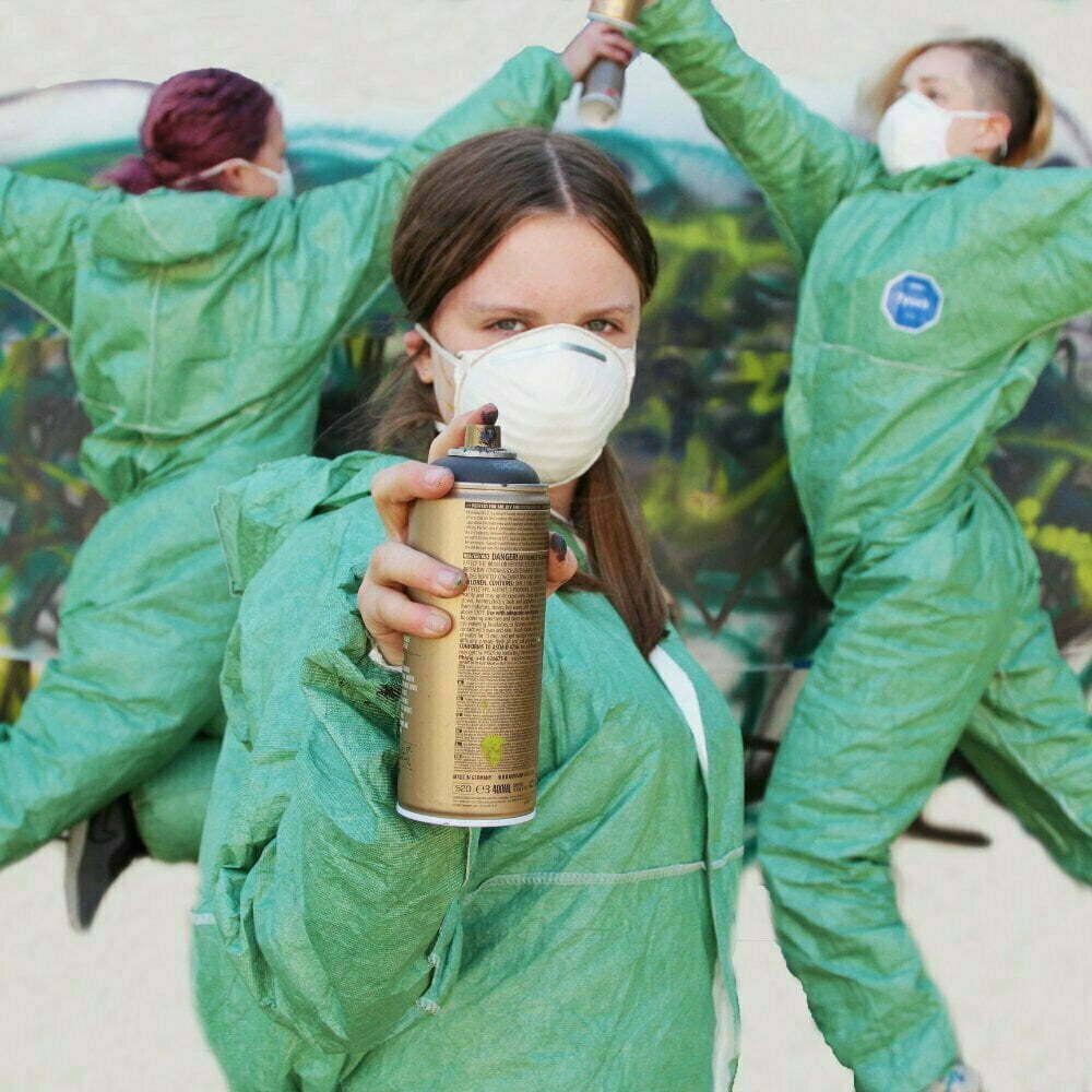 3 young dancers in green jumpsuits with facemasks and spray cans, dancing in front of graffiti on a wall