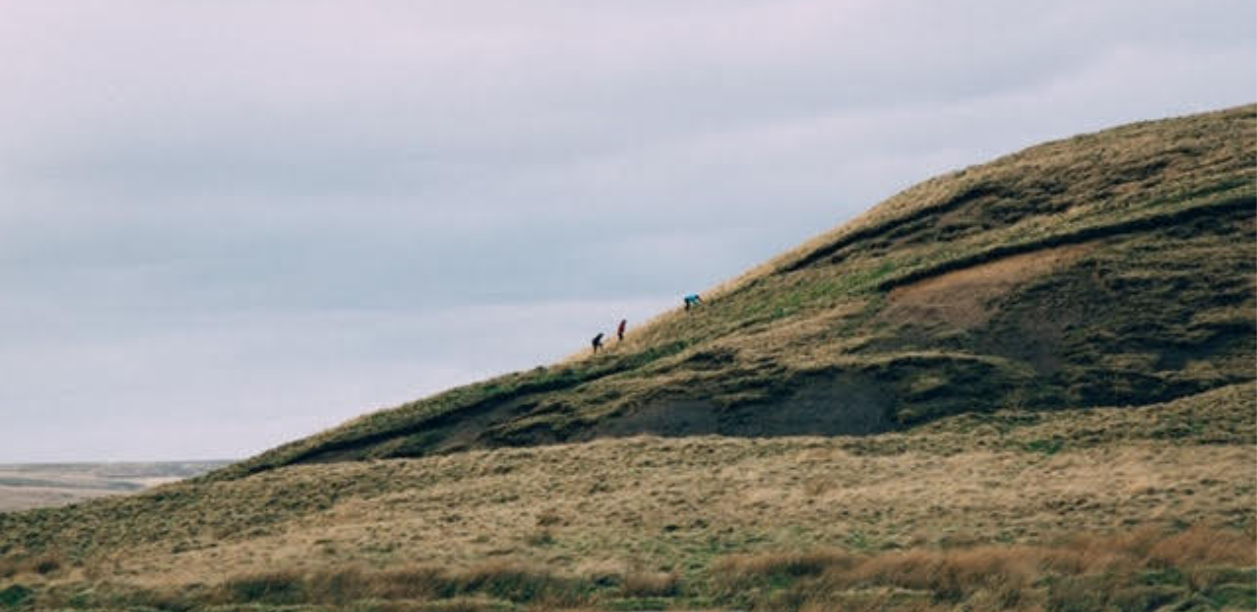 Viewed from a distance, three small figures are walking up a green hillside, which inclines upward from left to right. The figures are wearing bright waterproof jackets and are outlined against a grey, cloudy sky, which fills the upper left of the image.