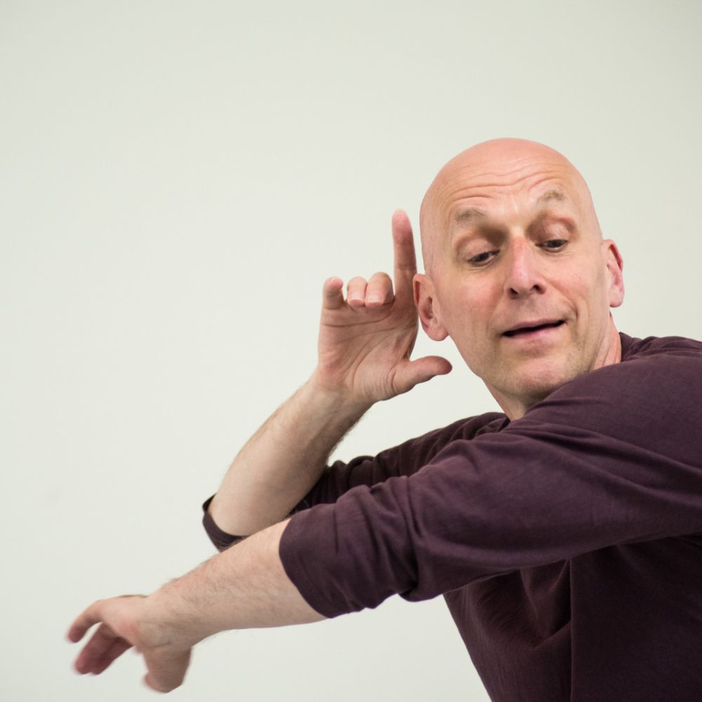 In close-up, a charismatic male dancer in a rich mulberry-coloured t-shirt poises his enfolding arms - one vertical and the other horizontal. He rests his right elbow on the back of his other hand and his right fingers nestle around his ear. Whatever music he is apparently hearing registers in a facial expression of inner bliss