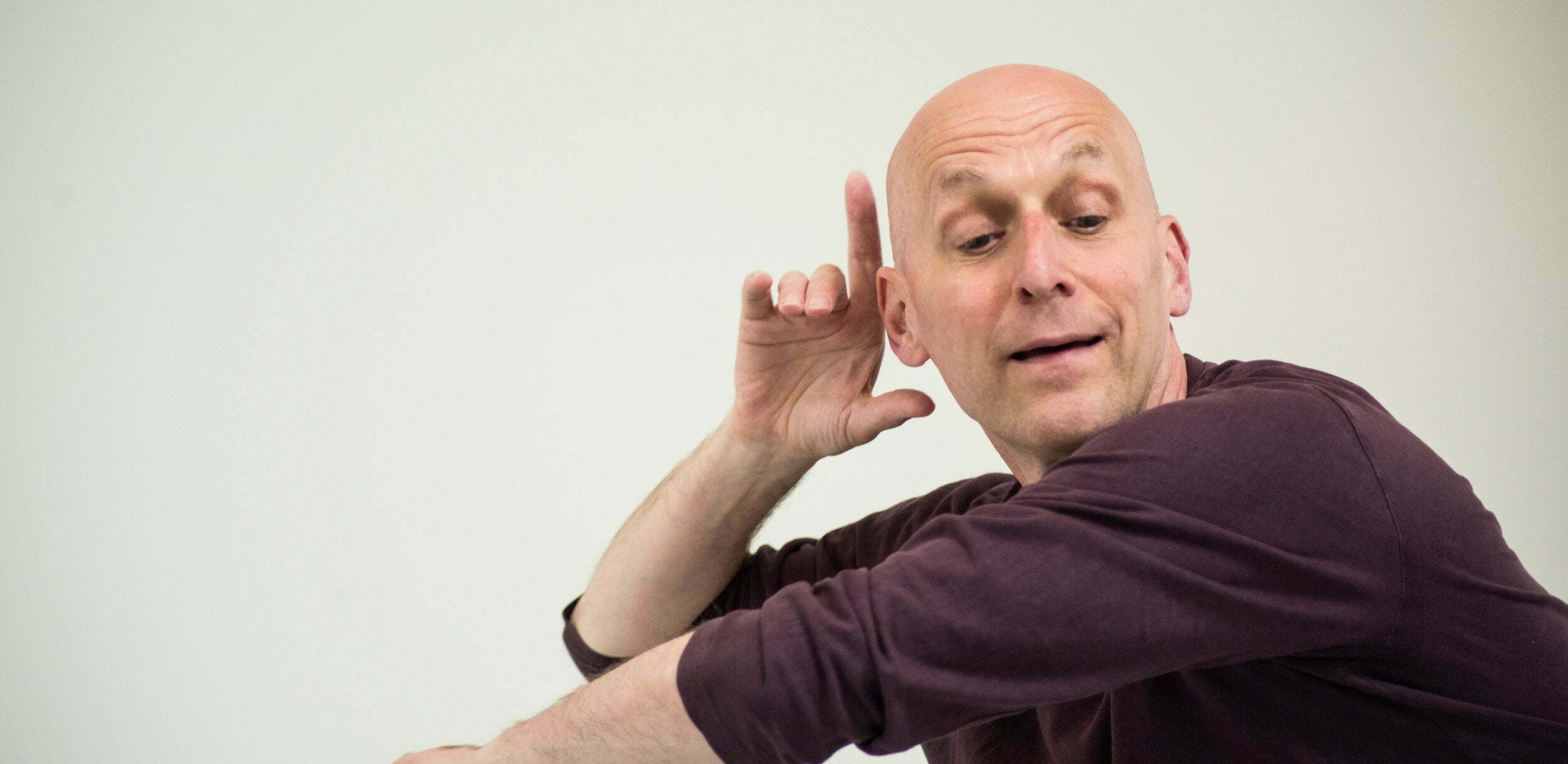 In close-up, a charismatic male dancer in a rich mulberry-coloured t-shirt poises his enfolding arms - one vertical and the other horizontal. He rests his right elbow on the back of his other hand and his right fingers nestle around his ear. Whatever music he is apparently hearing registers in a facial expression of inner bliss