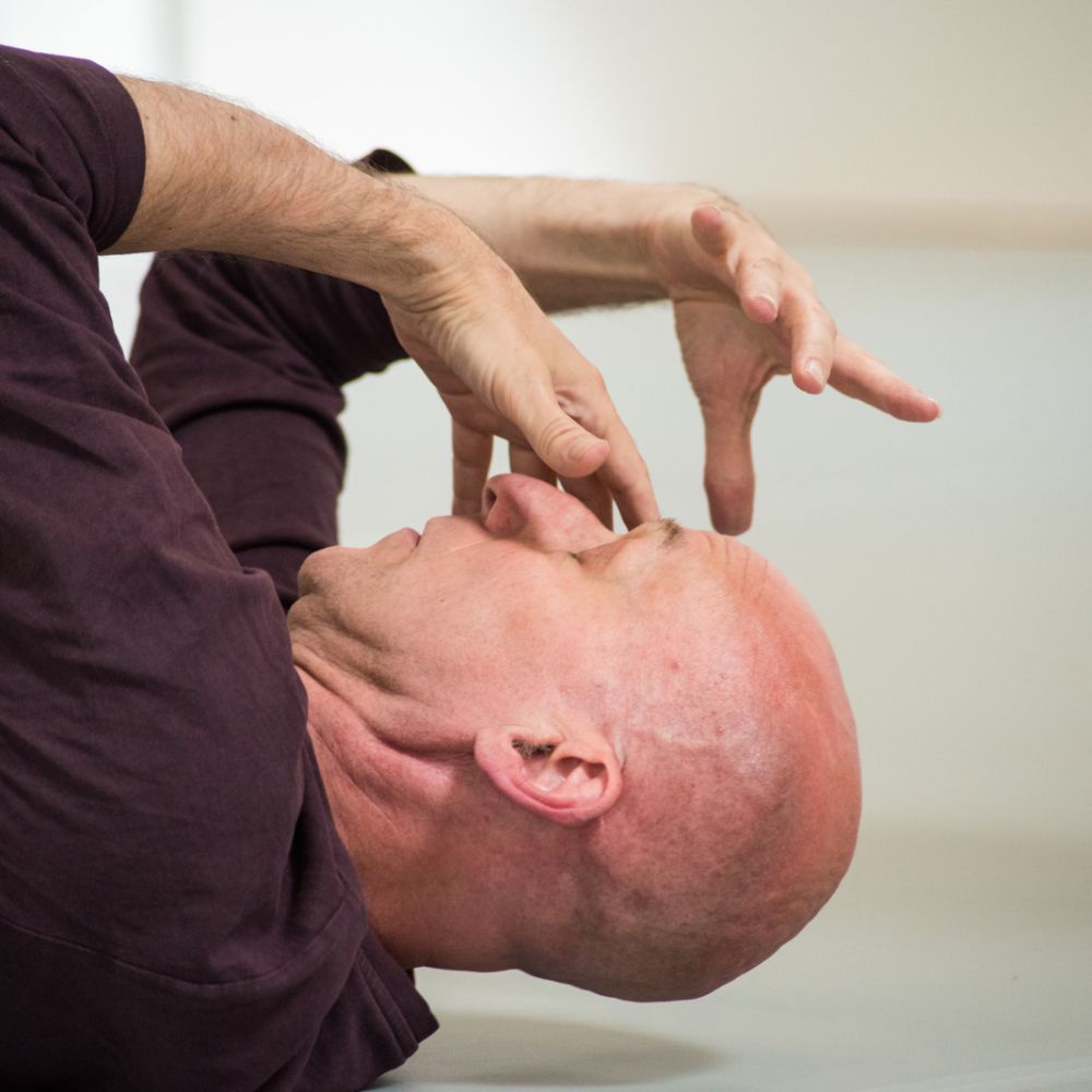 An intimate sculptural image of a dancer's bald head in profile. He's lying back on the ground, eyes closed. He seems to be having an intense experience - his fingers are scrunched and gnarly, poised mask-like in front of his face.
