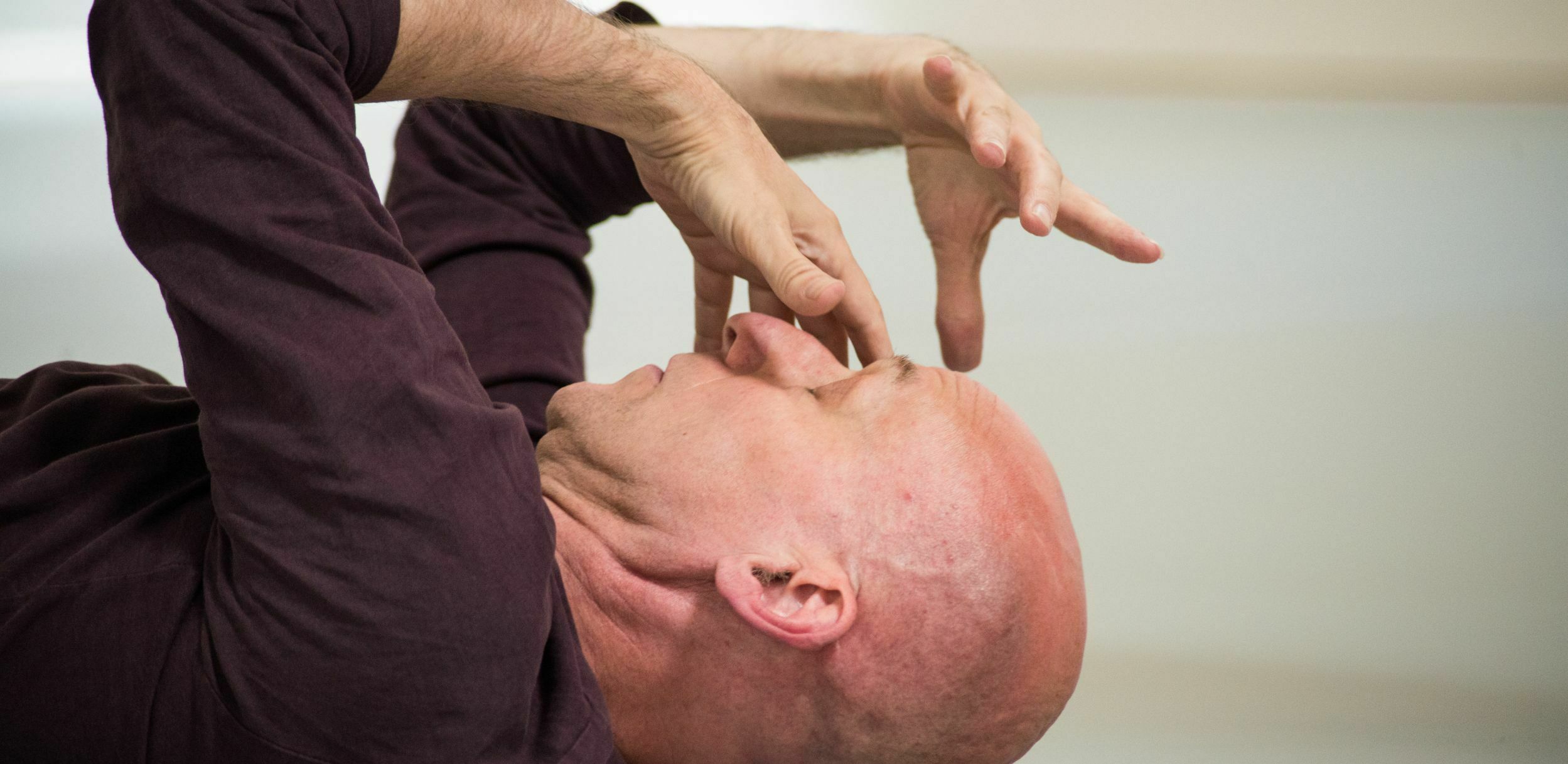 An intimate sculptural image of a dancer's bald head in profile. He's lying back on the ground, eyes closed. He seems to be having an intense experience - his fingers are scrunched and gnarly, poised mask-like in front of his face.